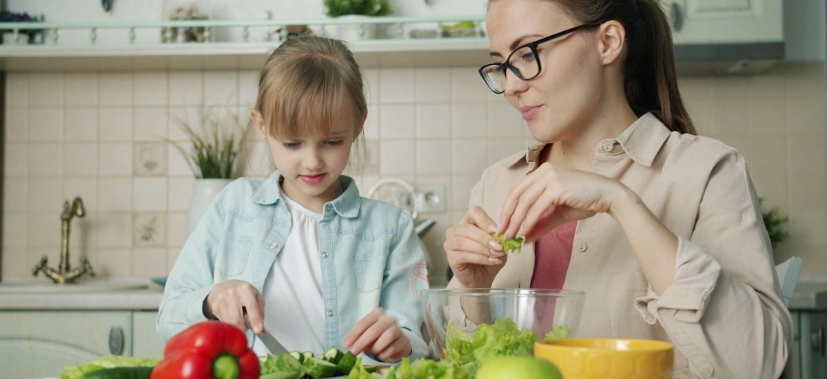 Cute loving daughter is helping mother with salad cutting cucumbers feeding young woman and laughing having fun at home. People and cookery concept.