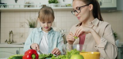 Cute loving daughter is helping mother with salad cutting cucumbers feeding young woman and laughing having fun at home. People and cookery concept.