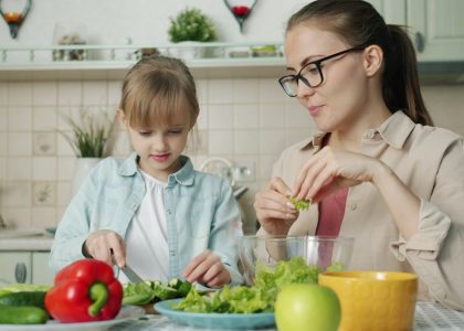 Cute loving daughter is helping mother with salad cutting cucumbers feeding young woman and laughing having fun at home. People and cookery concept.