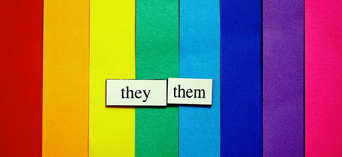 They/Them pronouns on rainbow-colored paper background.