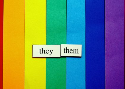 They/Them pronouns on rainbow-colored paper background.